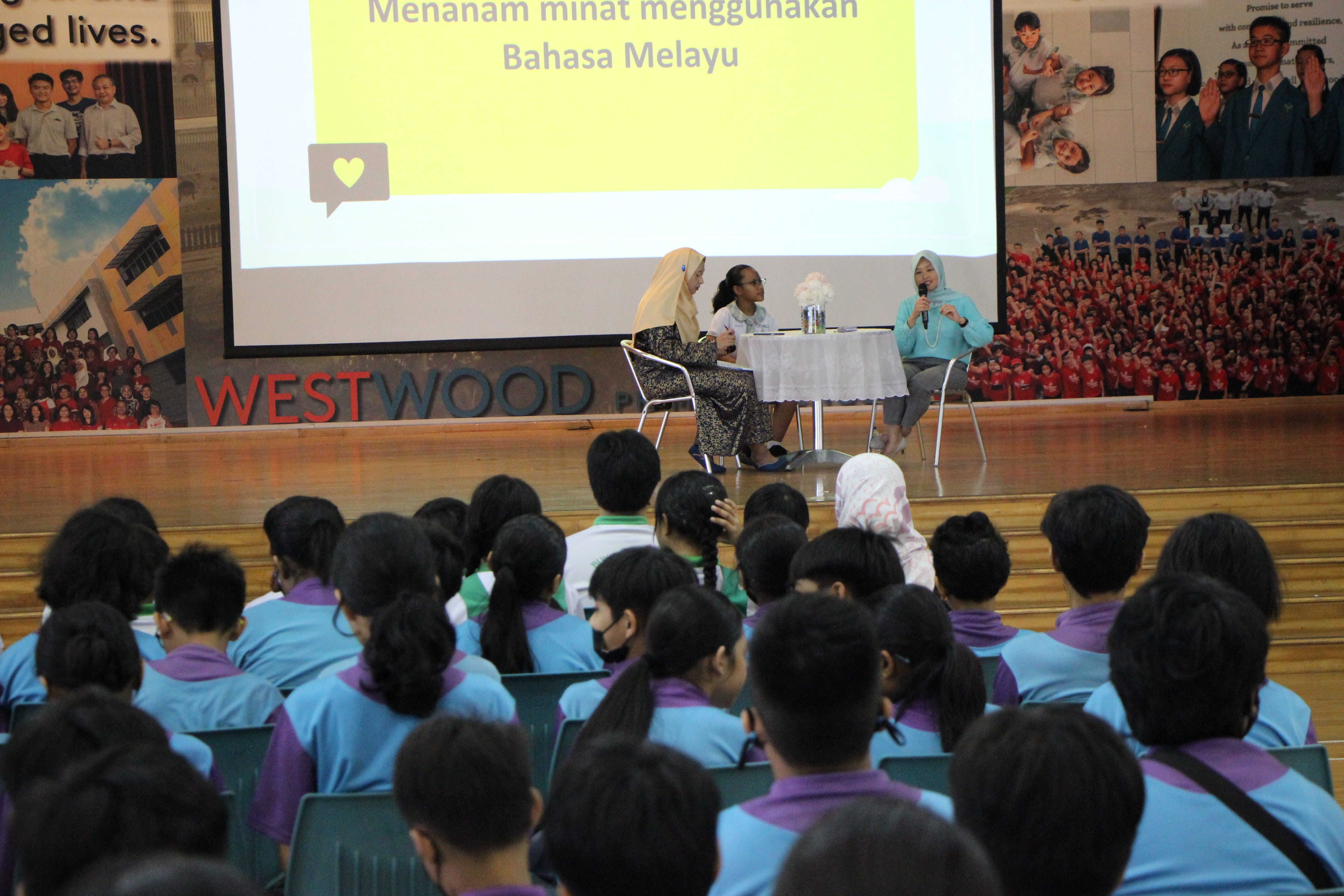 Three women on a Westwood school stage speak to a seated student audience under a Bahasa Melayu presentation slide.